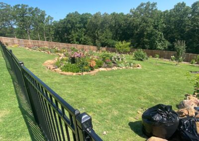Lush green lawn featuring a circular flower garden surrounded by a decorative stone border, with a black fence in the foreground, showcasing APEX Lawn Care's landscaping services in Little Rock, Arkansas.