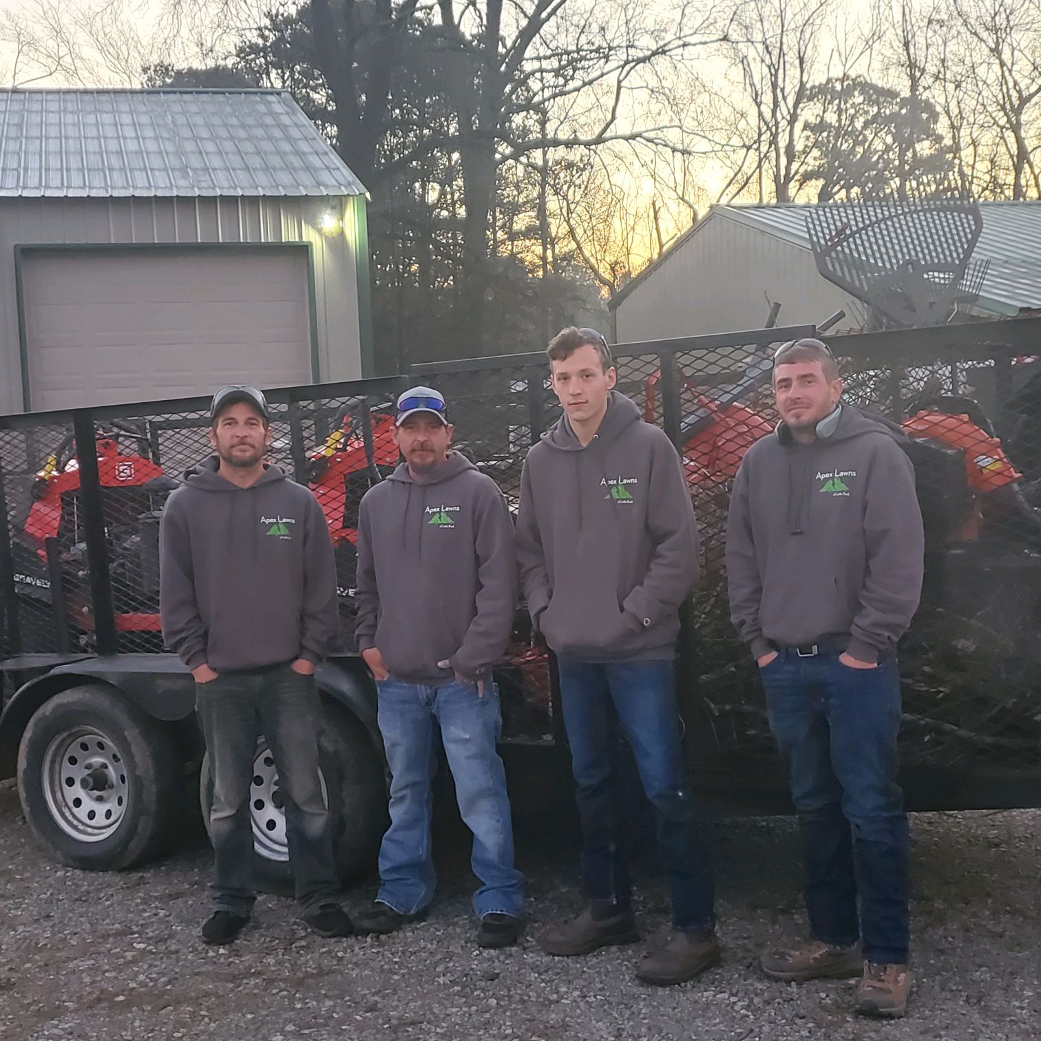 Four Apex Lawns employees in gray hoodies standing in front of a trailer filled with lawn maintenance equipment, showcasing their commitment to local lawn care services in Little Rock, Arkansas.
