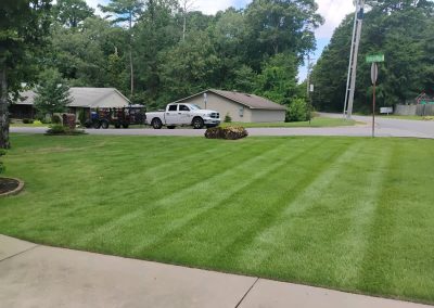 Lush green lawn with freshly mowed stripes, showcasing professional lawn care services by APEX Lawn Care in Central Arkansas, featuring a parked white truck and residential homes in the background.