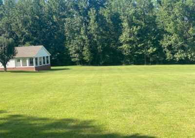 An employee in the response with the word employee and the Little Rock Metro Area a small white building with a red door adjacent to a mowed lawn and a line of trees in the background on a sunny day. - Little Rock Lawn Care and Mowing Services by Apex Lawn Care