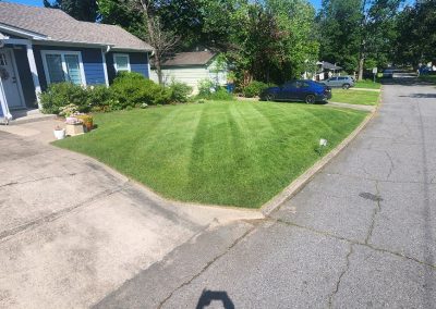 Lawn care service showcasing a well-maintained green lawn with striped patterns, bordered by a residential driveway and flower pots, highlighting APEX Lawn Care's professional landscaping in Central Arkansas.