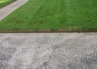 Lush green lawn with neatly trimmed grass adjacent to a cracked concrete driveway, showcasing APEX Lawn Care's professional lawn maintenance services in Central Arkansas.