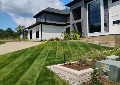 Lush green lawn with striped mowing patterns, landscaped garden beds, and modern home architecture in Central Arkansas, showcasing APEX Lawn Care's professional lawn care services.
